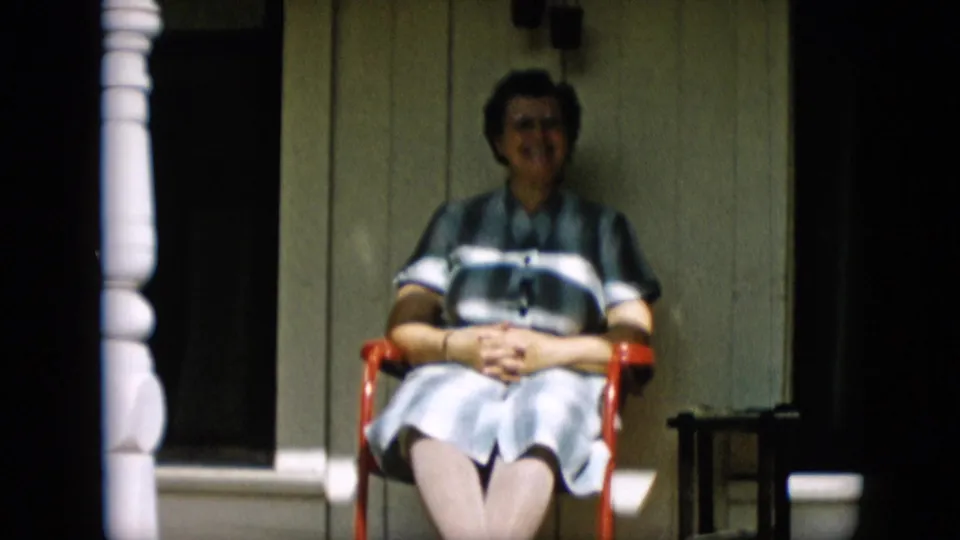 An older woman in a striped dress sits with hands clasped in a red chair on a porch, consistent with the 1957 Pennsylvania home-movie metadata.