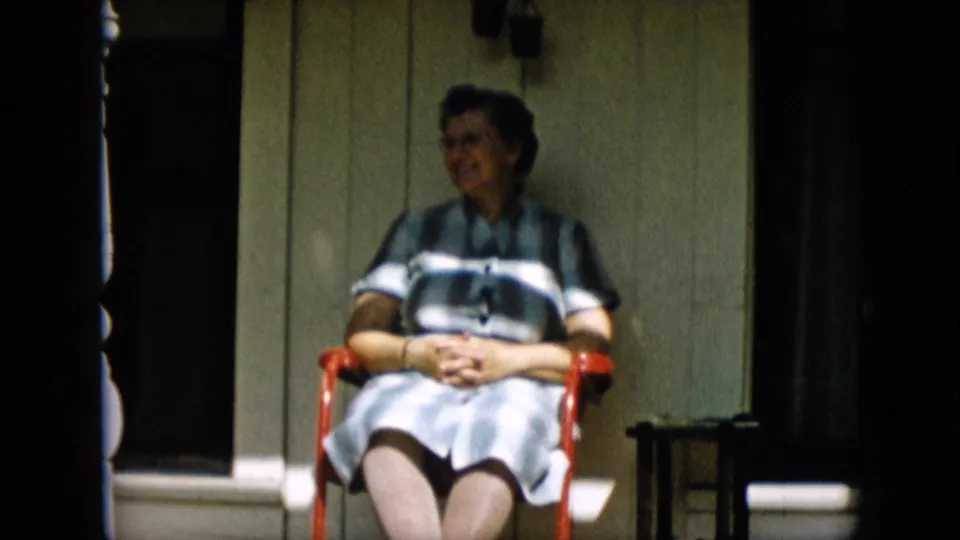 An older woman sits on a red porch chair with her hands folded, turning slightly while smiling in the 1957 Pennsylvania home-movie frame.