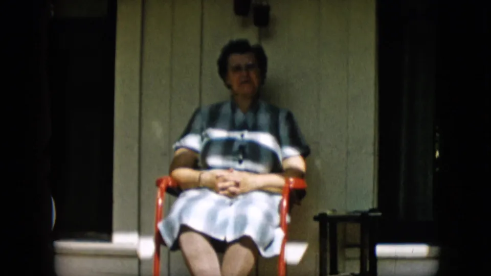 The woman faces the camera from a red chair on a shaded porch, with striped clothing, clasped hands, and white porch trim visible.