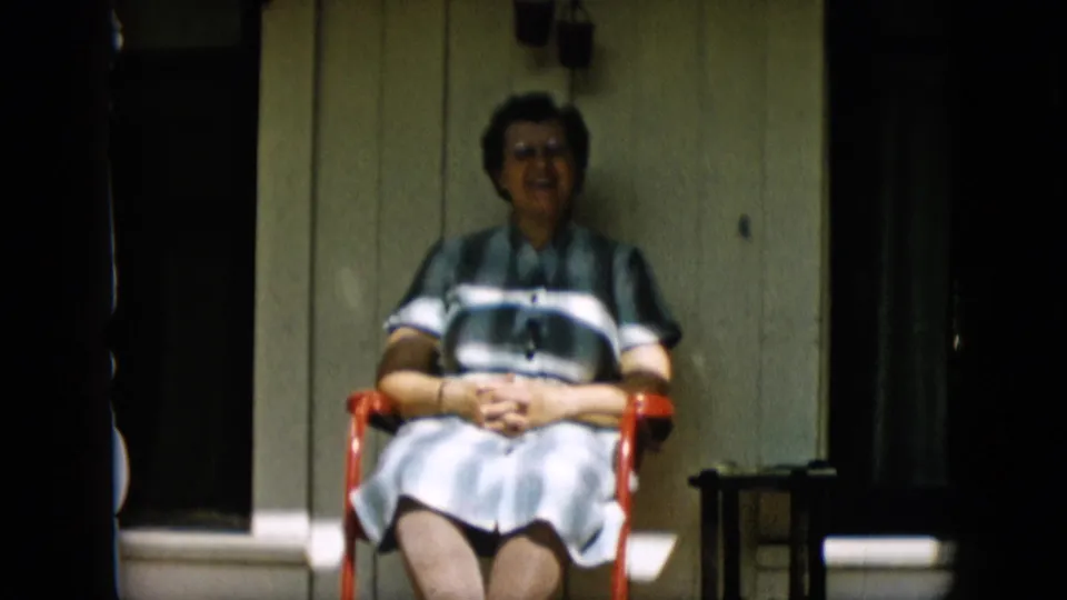 A smiling older woman in a striped dress remains seated in the red chair on a porch, with dark doorway shadows and white trim framing the scene.