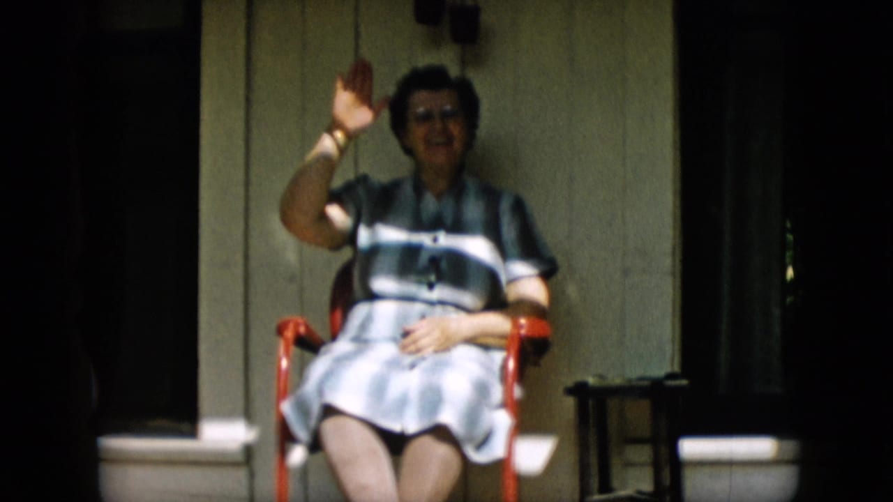 In supported 1957 Pennsylvania home-movie footage, an older woman in glasses and a striped dress sits on a porch in a red chair and begins waving toward the camera.