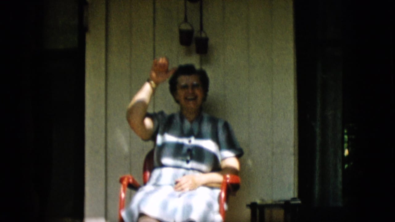 Older woman smiling from a red porch chair with one hand lifted in a wave.
