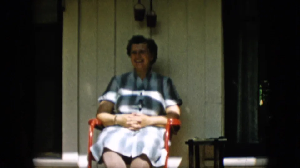 The woman sits back in the red porch chair with her hands clasped, framed by wood siding, shadowed doorways, and small hanging lanterns.