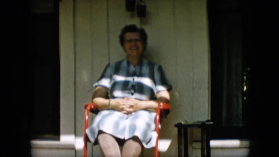 The 1957 Pennsylvania home-movie frame shows the older woman seated calmly on the porch, hands folded over her striped dress as sunlight falls across the chair and siding.