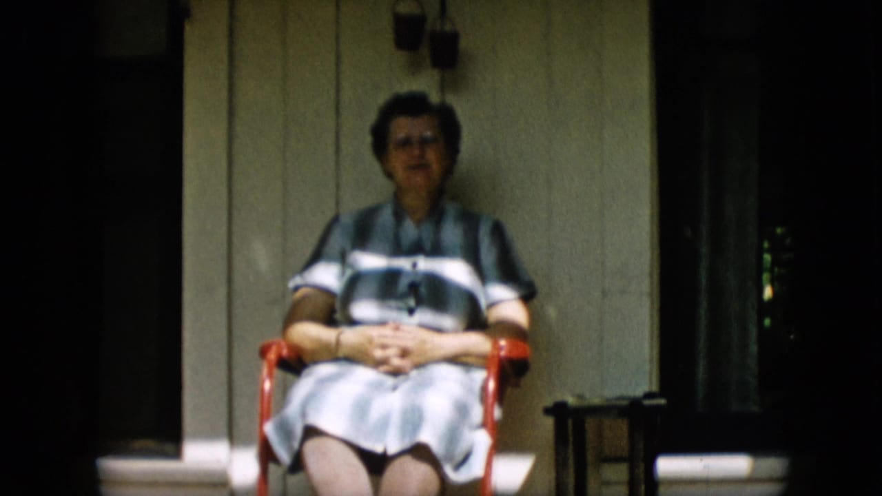 In this 1957 Pennsylvania home-movie frame, a woman wearing glasses and a striped dress sits on a red metal chair on a shaded porch with her hands clasped.