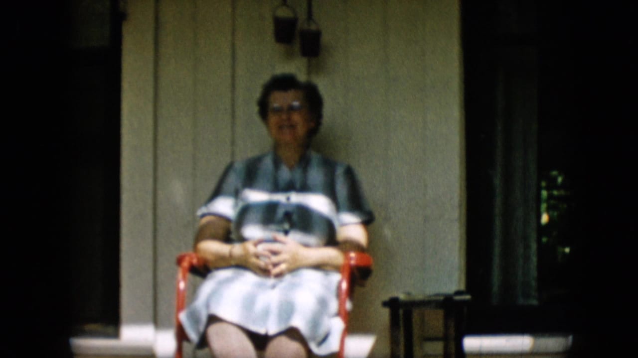 The woman remains seated on the red porch chair, facing the camera against light wooden siding with a dark doorway and small furniture nearby.