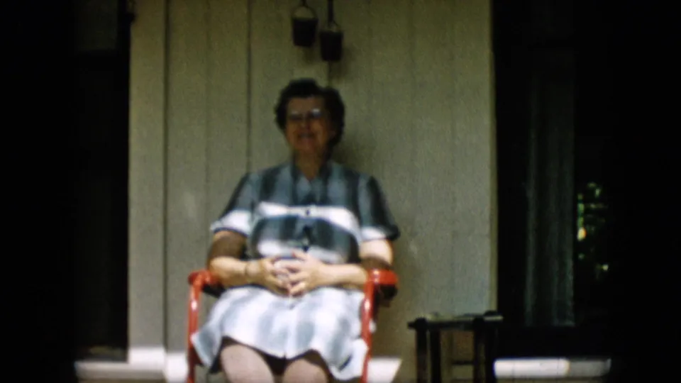 The woman remains seated on the red porch chair, facing the camera against light wooden siding with a dark doorway and small furniture nearby.