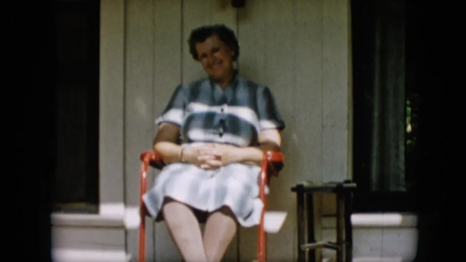 The woman tilts her head slightly while seated on the red chair, suggesting a conversational moment captured in the 1957 Pennsylvania home movie.