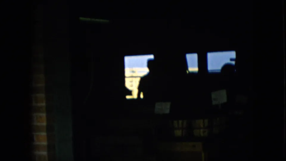 Pennsylvania 1957 8mm frame showing a dim building interior with a brick or wall edge, bright window openings, small signs, and indistinct silhouetted shapes.
