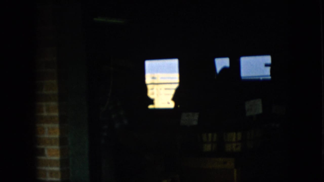 1957 Pennsylvania 8mm frame showing a very dark indoor market or shop area with bright window-like rectangles, a brick edge, and a partially visible person in plaid shadow.
