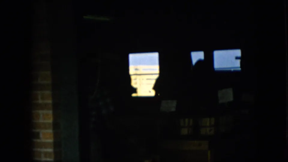 1957 Pennsylvania 8mm frame showing a very dark indoor market or shop area with bright window-like rectangles, a brick edge, and a partially visible person in plaid shadow.