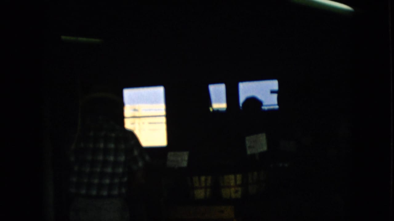 1957 Pennsylvania 8mm frame showing a very dark home interior with a person in a checkered shirt near bright rectangular windows and other indistinct figures in shadow.