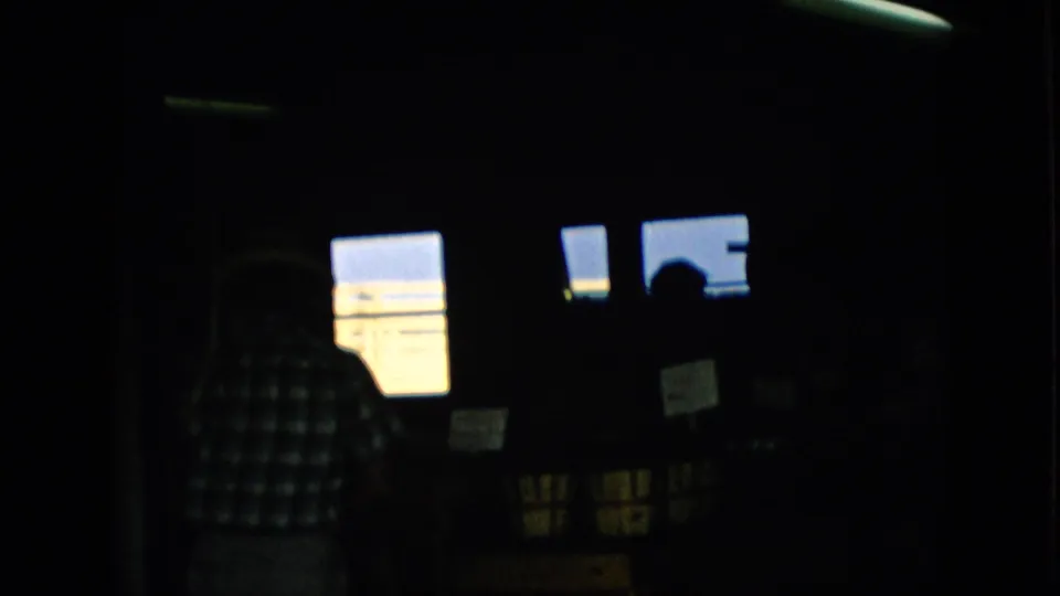 1957 Pennsylvania 8mm frame showing a very dark home interior with a person in a checkered shirt near bright rectangular windows and other indistinct figures in shadow.