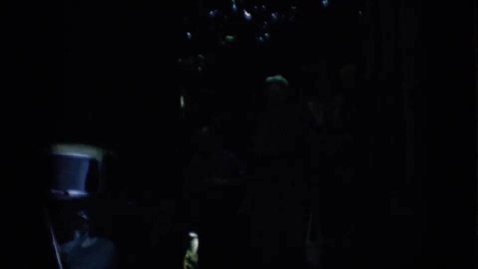 Dark 8mm frame from the 1957 Pennsylvania clip showing small reflected lights, a chair at the lower left, and indistinct human shapes in deep shadow.