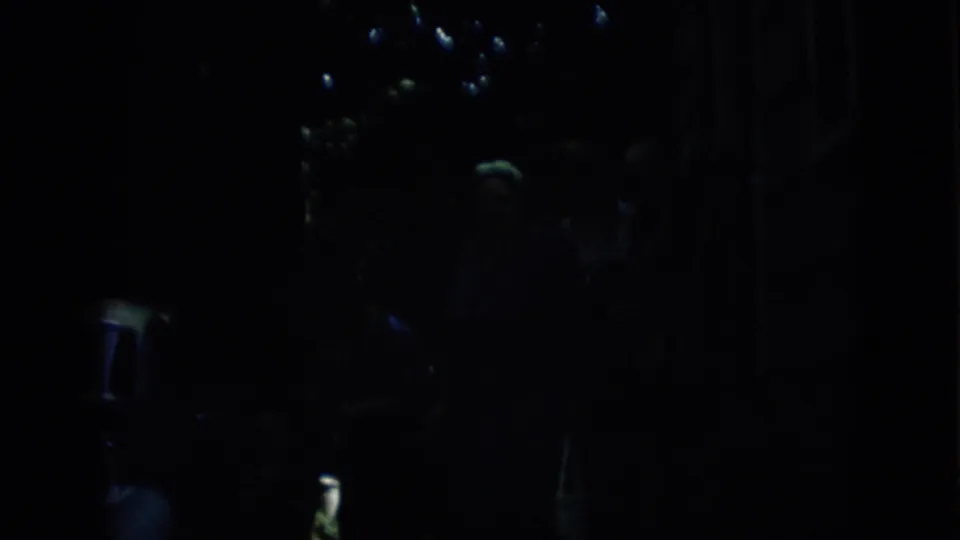 1957 Pennsylvania 8mm frame showing a nearly black nighttime view with faint points of light, indistinct figures, and a small illuminated patch of foliage at the upper right.