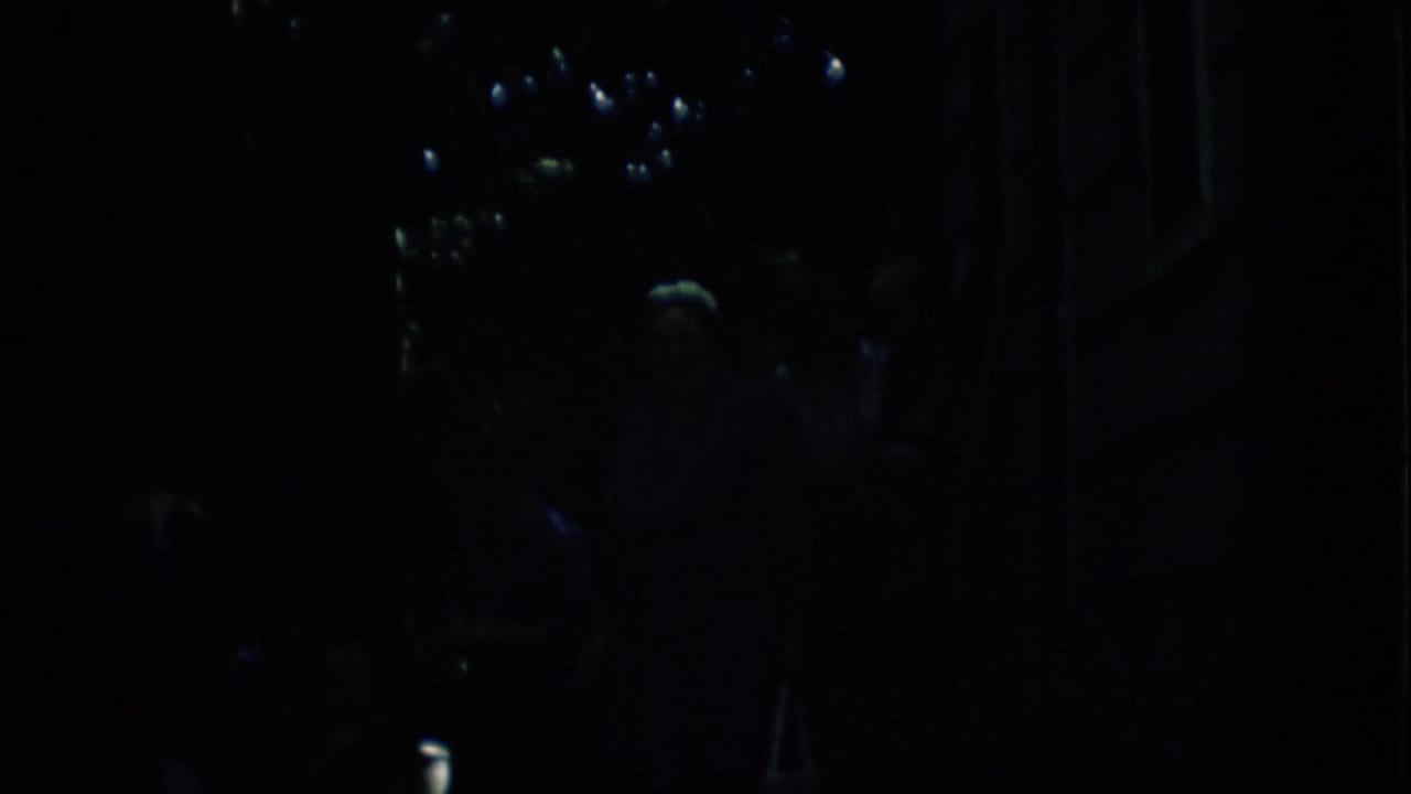 Extremely low-light 1957 Pennsylvania 8mm frame showing indistinct figures near a dark street-side area with small reflected lights and deep shadows.