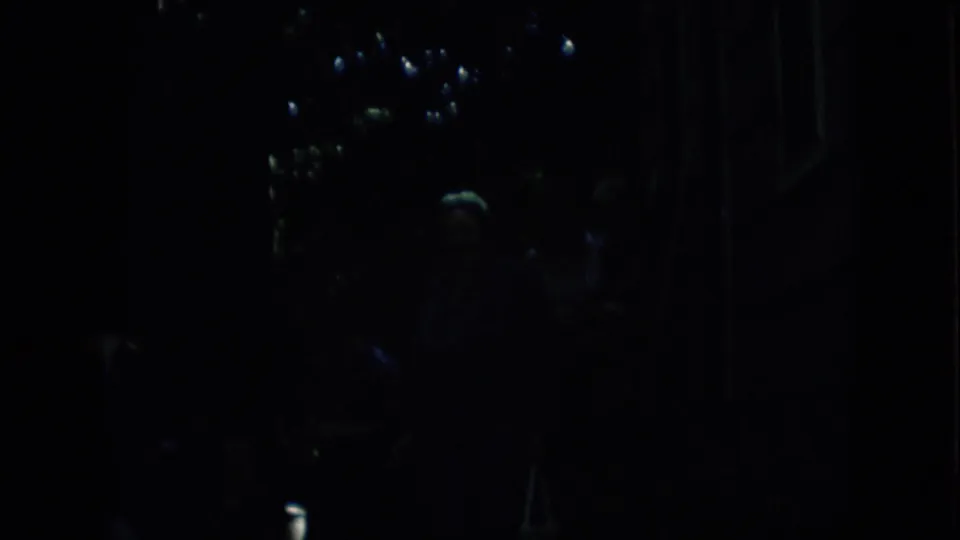 Extremely low-light 1957 Pennsylvania 8mm frame showing indistinct figures near a dark street-side area with small reflected lights and deep shadows.