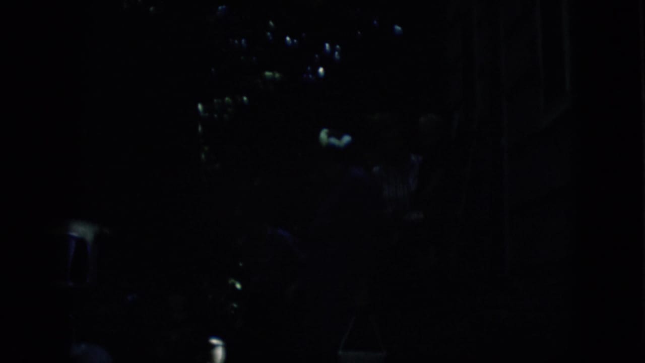 Dark nighttime frame with faint figures, small highlights, and a shadowed object that may be a car or street fixture.