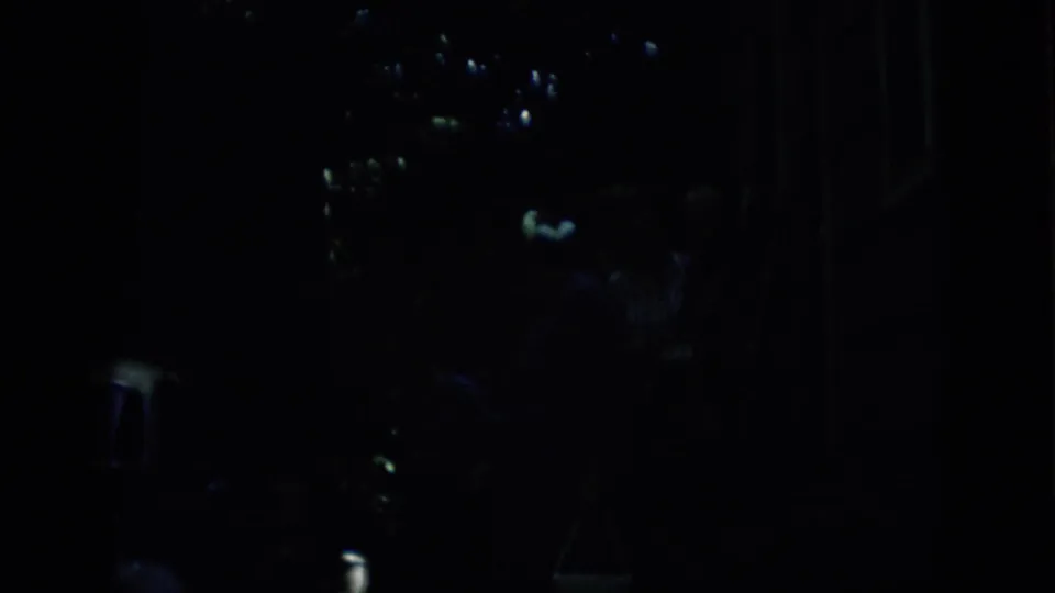 Low-exposure 1957 Pennsylvania home-movie frame with faint human figures near scattered highlights and a possible parked car or street-side object.