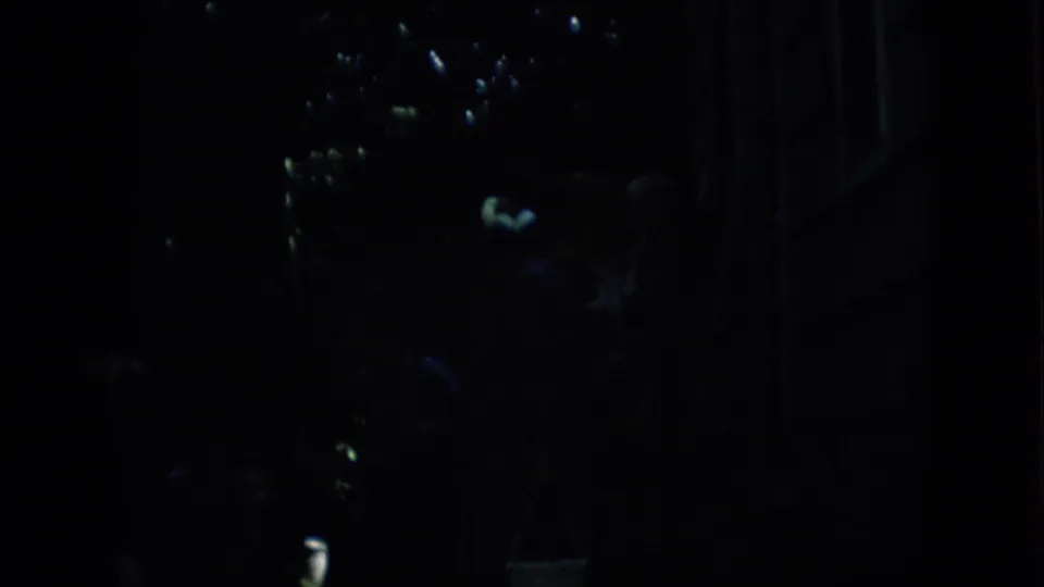 Dark 1957 Pennsylvania 8mm frame showing several hard-to-identify people near a bright highlight and faint stair or building-edge shapes.