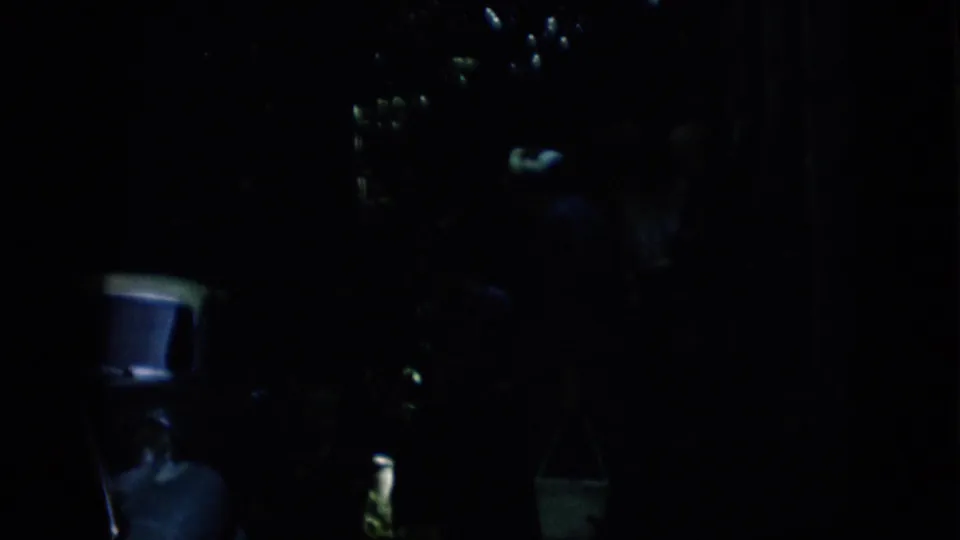 Pennsylvania 1957 home-movie frame showing part of a light-colored automobile in heavy shade, with a hanging white bucket and faint yard detail nearby.