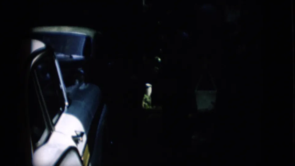 Pennsylvania 1957 home-movie frame showing a closer view of the car's side and window area, with a dim figure working near the vehicle and a hanging bucket in the background.