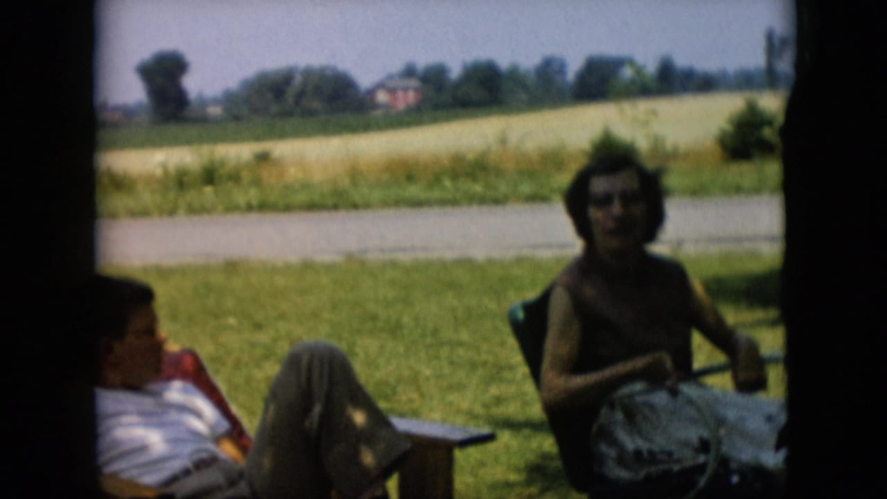 1957 Pennsylvania 8mm frame showing a woman seated in a lawn chair and a man reclining nearby, with grass, a road, fields, and distant trees behind them.