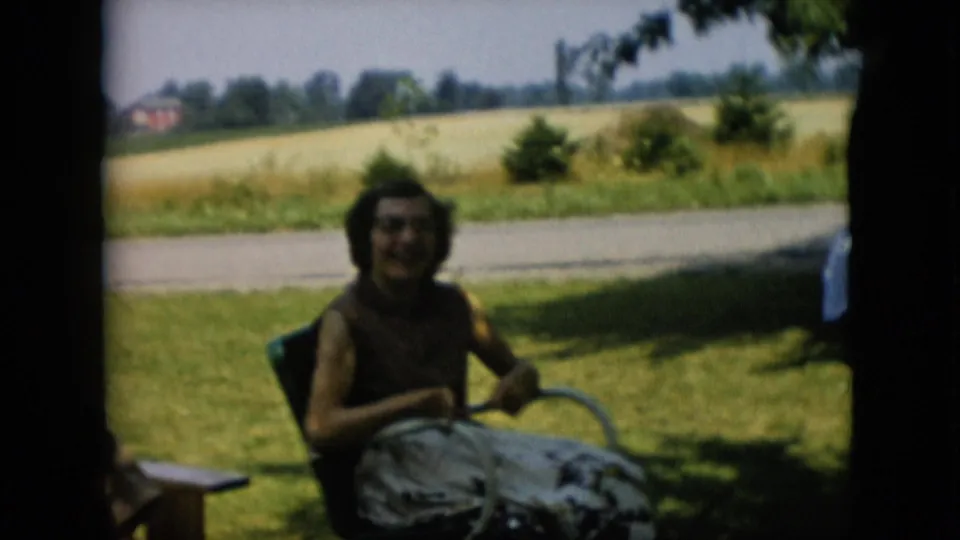 1957 Pennsylvania 8mm frame showing a woman laughing from a lawn chair in a grassy yard, with open fields and a distant building beyond the road.