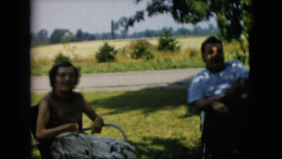 1957 Pennsylvania home-movie frame with a woman seated at center and a man in a striped shirt at right, both relaxing in a shaded yard near open fields.
