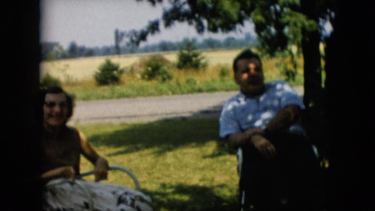 1957 Pennsylvania 8mm home-movie frame showing a man and woman seated in lawn chairs under tree shade near a road and open field.