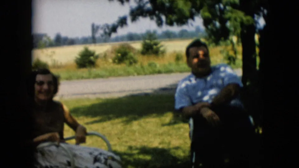 1957 Pennsylvania 8mm home-movie frame showing a man and woman seated in lawn chairs under tree shade near a road and open field.