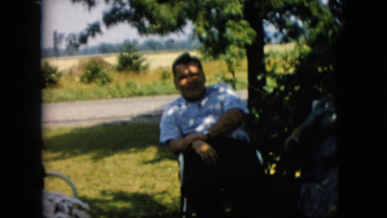A man sits in shade under a tree with a road and field behind him.