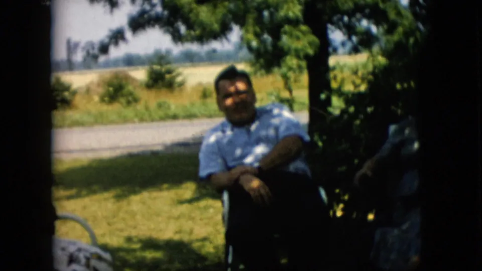 1957 Pennsylvania frame with a man in a light patterned shirt seated under tree shade near a roadside field, with another person partly visible at the edge.