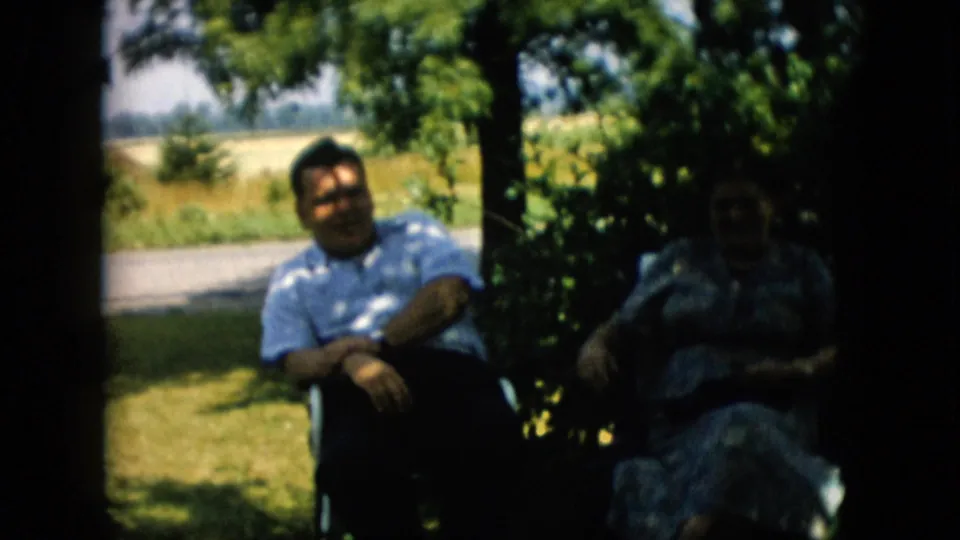 1957 Pennsylvania 8mm frame showing two adults in lawn chairs beneath tree shade, with the man clearly visible and the woman partly obscured by shadow.