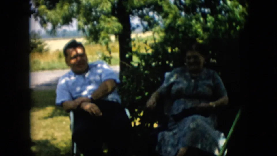 1957 Pennsylvania home-movie frame of a man and woman seated under dense tree shade, with grass, a roadside, and open summer fields in the background.