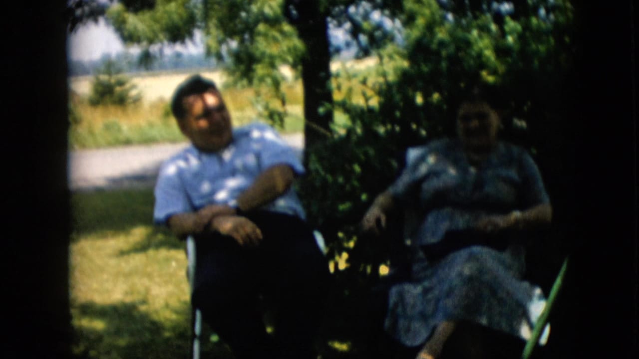 In 1957 Pennsylvania home-movie footage, a man in a light patterned shirt and a woman in a floral dress sit in lawn chairs under leafy shade near an open yard.