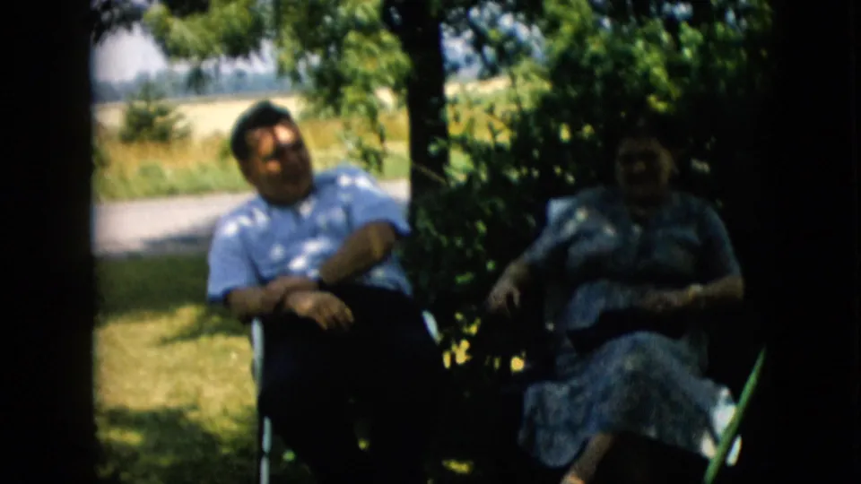In 1957 Pennsylvania home-movie footage, a man in a light patterned shirt and a woman in a floral dress sit in lawn chairs under leafy shade near an open yard.
