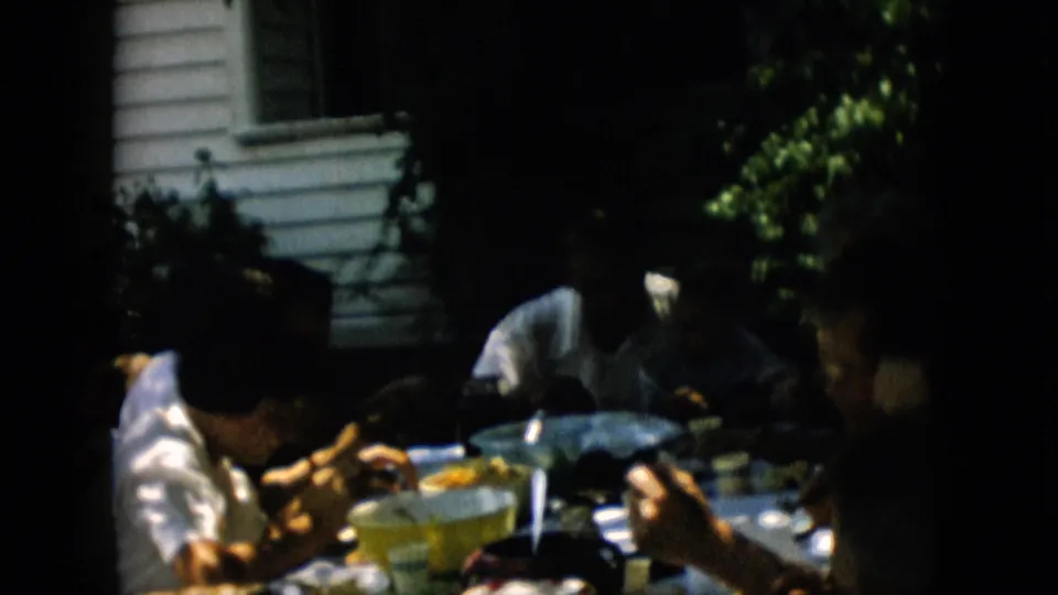 A shaded outdoor dining scene shows several family members seated close together at a table with food, bowls, plates, and utensils visible.
