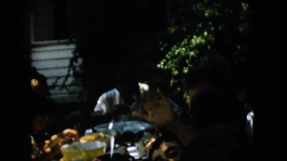 The frame shows a close, darkly shaded view of family members eating at an outdoor table with bowls, plates, and a house exterior partly visible.
