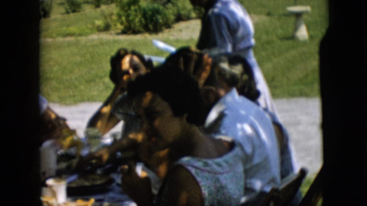 1957 Pennsylvania home-movie frame showing several adults seated at a shaded picnic table with plates, cups, bottles, and a sunlit lawn behind them.