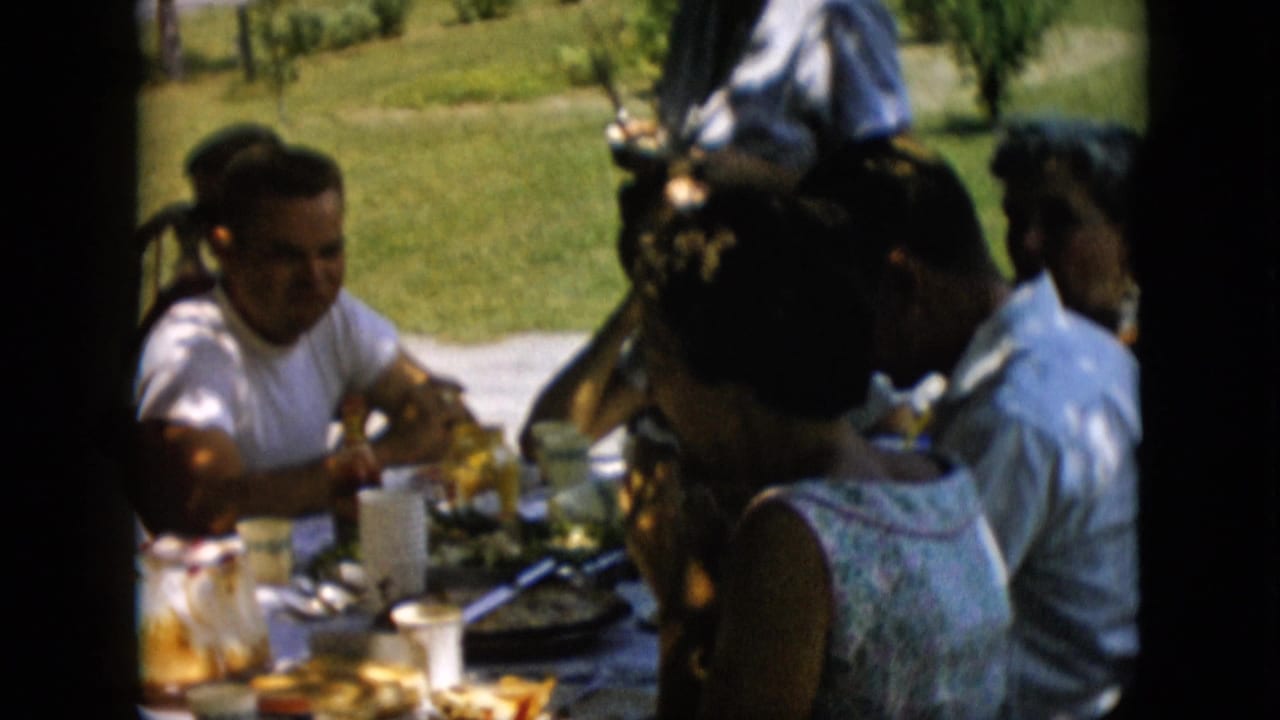 1957 Pennsylvania frame showing a woman in a white shirt seated at an outdoor picnic table with other adults partly visible around food and drinks.