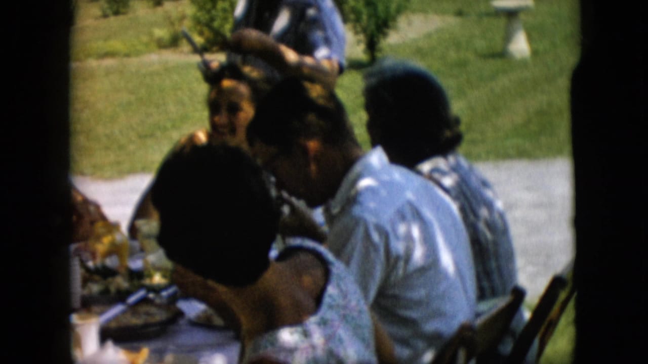 Adults sit close together at a picnic table under shade with a lawn and path behind them.