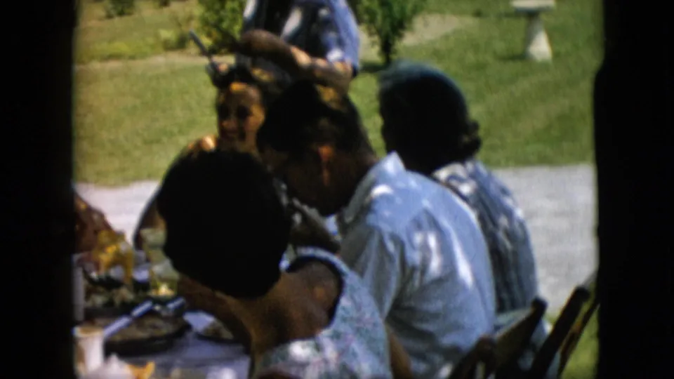 1957 Pennsylvania home-movie frame of adults seated closely at a backyard picnic table, with strong sunlight on the lawn and a paved path in the background.