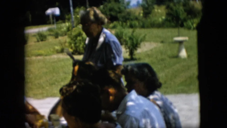 1957 Pennsylvania frame showing a standing adult beside a shaded picnic table, with seated adults, a green lawn, shrubs, path, and stone bird bath visible behind them.
