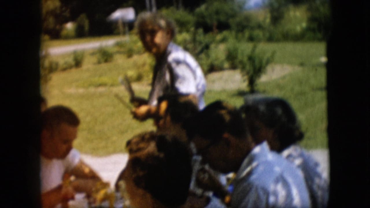 In a 1957 Pennsylvania home-movie frame, adults sit around an outdoor picnic table on a grassy lawn while one man stands in the background.