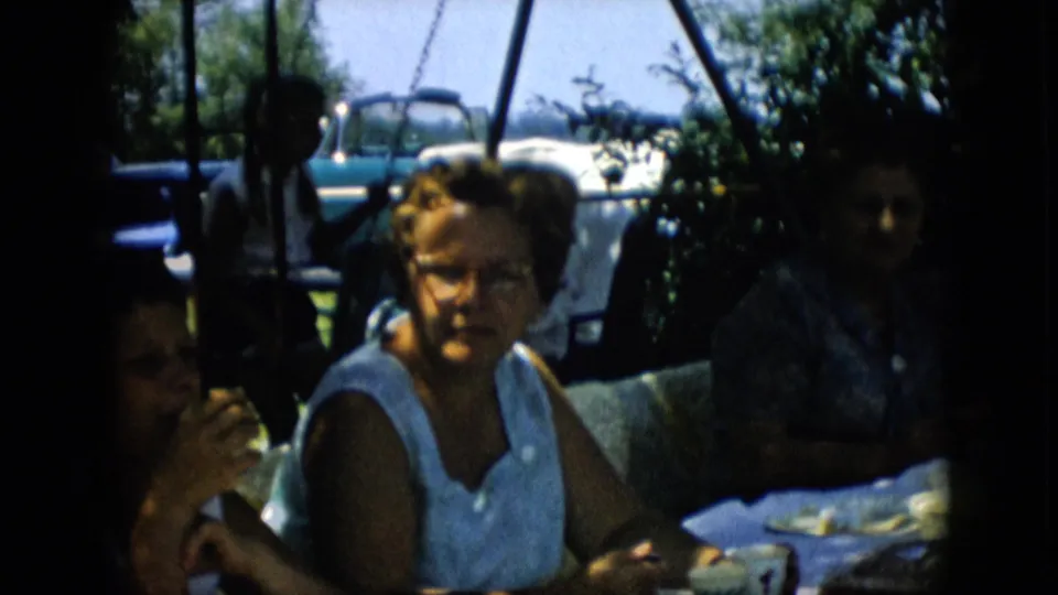 A woman in a white sleeveless top sits near the picnic table in the 1957 Pennsylvania home movie, with a swing set and parked car visible in the yard behind her.