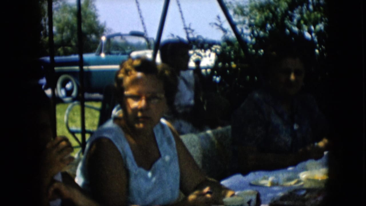 1957 Pennsylvania home-movie frame showing adults seated at a shaded picnic table, with a woman in a light sleeveless dress in front and a blue-and-white convertible parked behind them.