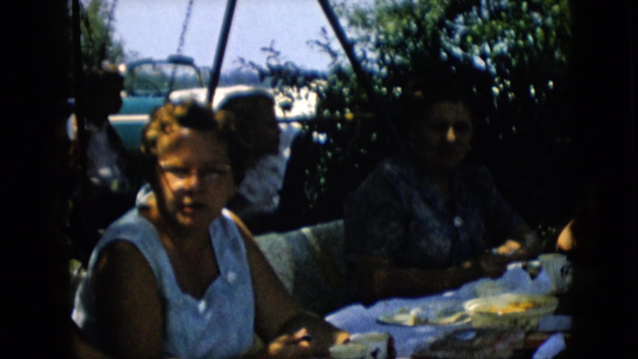 Two women and other adults sit at an outdoor table with dishes and a convertible visible behind greenery.