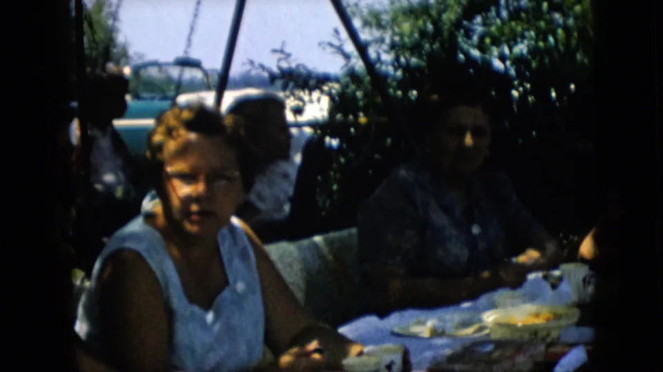 1957 Pennsylvania frame of a family group seated outdoors at a picnic table, including a woman in glasses in the foreground and a patterned-blouse woman partly in shadow.