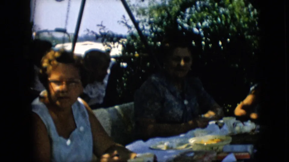 1957 Pennsylvania home-movie frame with the foreground woman partly cropped, other adults seated at the picnic table, and a classic convertible still visible beyond the yard.